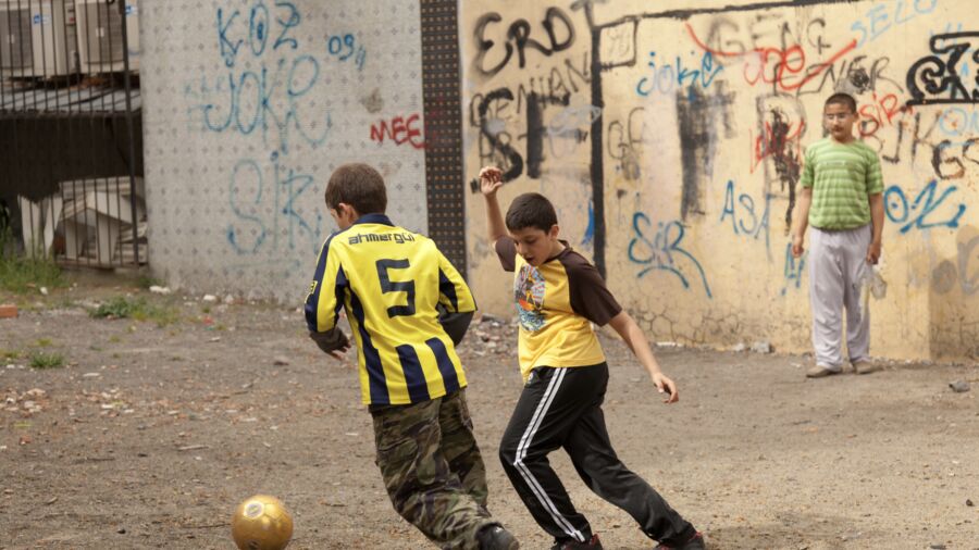 Three children playing soccer on a dirt field.