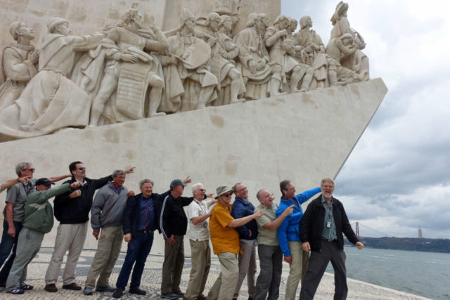 A line of men excitedly pointing forward, with Rick Steves at the front smiling and facing the camera, in front of a large stone monument that looks similar